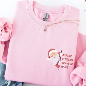 Op de afbeelding: Een lichtroze sweatshirt met een ronde hals. Het sweatshirt heeft een afbeelding van de kerstman met een bord met de tekst "Judging Behaviors Including Yours". Een gouden ketting met een kleine hanger is over het sweatshirt gedrapeerd.