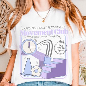 May include: White t-shirt with a graphic design featuring the text "UNAPOLOGETICALLY PLAY-BASED Movement Club Building Strength Through Play." The design includes a stopwatch, stairs, a soccer ball, and the phrase "Small Wins. Big Progress."