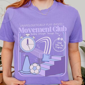 May include: A lilac-colored t-shirt with a graphic design featuring a stopwatch, soccer ball, cone, and stairs. The text on the shirt reads "UNAPOLOGETICALLY PLAY-BASED Movement Club" and "Building Strength Through Play". The design also includes the phrase "Small Wins. Big Progress."