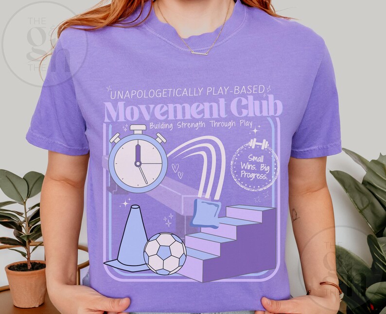 May include: A lilac-colored t-shirt with a graphic design featuring a stopwatch, soccer ball, cone, and stairs. The text on the shirt reads "UNAPOLOGETICALLY PLAY-BASED Movement Club" and "Building Strength Through Play". The design also includes the phrase "Small Wins. Big Progress."