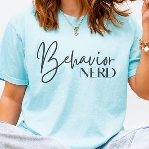 Puede incluir: Camiseta azul claro con la frase "Behavior Nerd" en negro, con letras cursivas y de bloque. La camiseta tiene mangas cortas y un corte holgado, ideal para el uso diario.