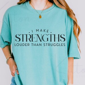May include: A turquoise t-shirt with the black text "I MAKE STRENGTHS LOUDER THAN STRUGGLES". The shirt has a crew neck and short sleeves. A gold necklace is worn.