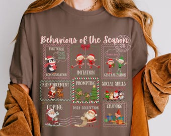 May include: A brown t-shirt with a festive graphic design. The design features the text "Behaviors of the Season" and various Christmas-themed illustrations, including stamps with words such as "Communication" and "Imitation."