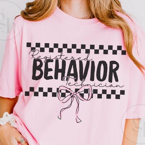 以下が含まれることがあります： 「Registered Behavior Technician」の黒い文字とリボン、白黒のチェック柄が特徴の、明るいピンク色のTシャツ。クルーネックのスタイルです。