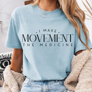 May include: Light blue t-shirt with the text "I MAKE MOVEMENT THE MEDICINE" in black. The shirt is a soft, casual style. The model is wearing a necklace and jeans.
