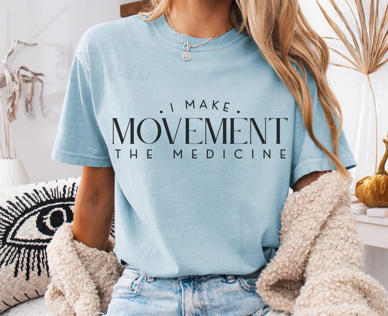 May include: Light blue t-shirt with the text "I MAKE MOVEMENT THE MEDICINE" in black. The shirt is a soft, casual style. The model is wearing a necklace and jeans.