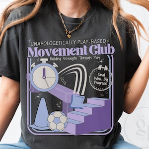 May include: Dark gray t-shirt with a graphic design featuring the text "UNAPOLOGETICALLY PLAY-BASED Movement Club" and illustrations of a stopwatch, stairs, a soccer ball, and a cone. The shirt also includes the phrase "Small Wins. Big Progress."
