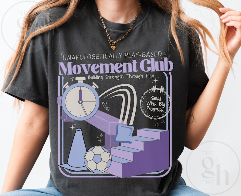 May include: Dark gray t-shirt with a graphic design featuring the text "UNAPOLOGETICALLY PLAY-BASED Movement Club" and illustrations of a stopwatch, stairs, a soccer ball, and a cone. The shirt also includes the phrase "Small Wins. Big Progress."