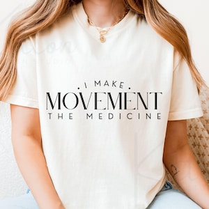 May include: Cream-colored t-shirt with the text "I MAKE MOVEMENT THE MEDICINE" in black. The shirt has a crew neck and short sleeves. The image shows the shirt on a person.