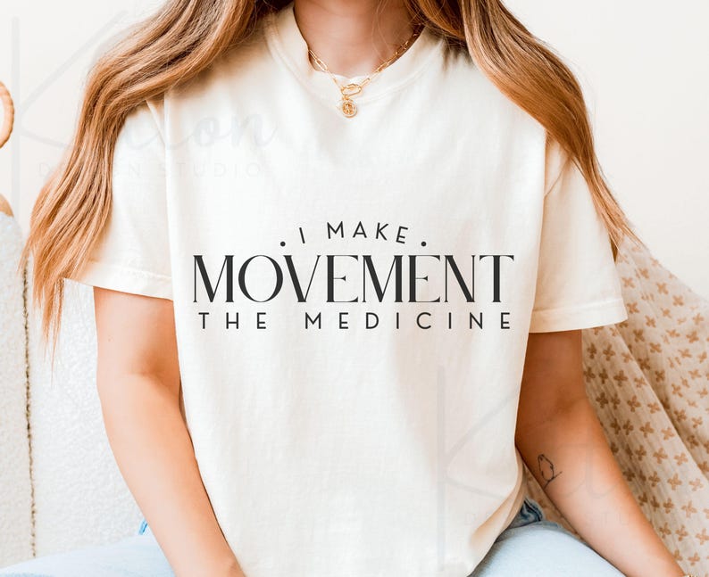 May include: Cream-colored t-shirt with the text "I MAKE MOVEMENT THE MEDICINE" in black. The shirt has a crew neck and short sleeves. The image shows the shirt on a person.