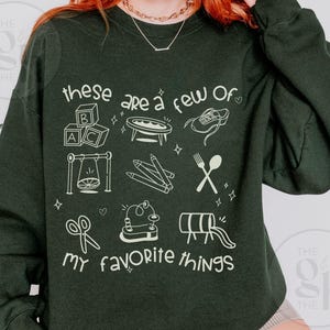 May include: Dark green sweatshirt with the text "these are a few of my favorite things" in white. The design includes line drawings of various objects, such as ABC blocks, a swing, pencils, and a shoe.