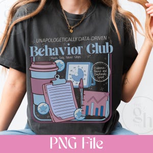 Op de afbeelding: Donkergrijs t-shirt met een grafisch ontwerp met de tekst "Behavior Club" en ondersteunende afbeeldingen. Het ontwerp bevat een koffiekopje, een klembord, een klok en een staafdiagram. De tekst "Unapologetically Data-Driven" staat boven de hoofdtekst.