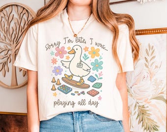 May include: Cream-colored t-shirt with a playful graphic of a duck on a skateboard, surrounded by colorful flowers and game pieces. The text reads "Sorry I'm late, I was... playing all day."