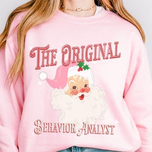 May include: Pink sweatshirt with the text "The Original Behavior Analyst" in red script. The design features a vintage Santa Claus illustration with a pink hat and white beard. A festive and humorous design.
