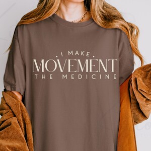 May include: A brown t-shirt with the text "I MAKE MOVEMENT THE MEDICINE" in a cream-colored, serif font. The shirt is worn by a person with long, wavy hair and a brown jacket.