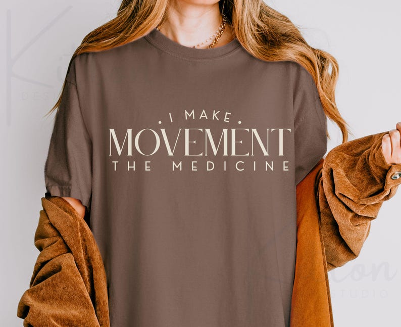 May include: A brown t-shirt with the text "I MAKE MOVEMENT THE MEDICINE" in a cream-colored, serif font. The shirt is worn by a person with long, wavy hair and a brown jacket.