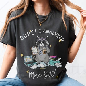 以下が含まれることがあります： アライグマのグラフィックがプリントされた濃いグレーのTシャツ。アライグマはメガネをかけ、クリップボードとコーヒーを持っています。シャツのテキストは「Oops! I Analyzed More Data!」で、ラップトップ、プリンター、本が描かれています。