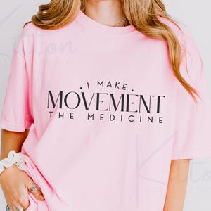 May include: A light pink t-shirt with the text "I MAKE MOVEMENT THE MEDICINE" in black. The shirt has short sleeves and a relaxed fit. The image is a close-up shot of the shirt.