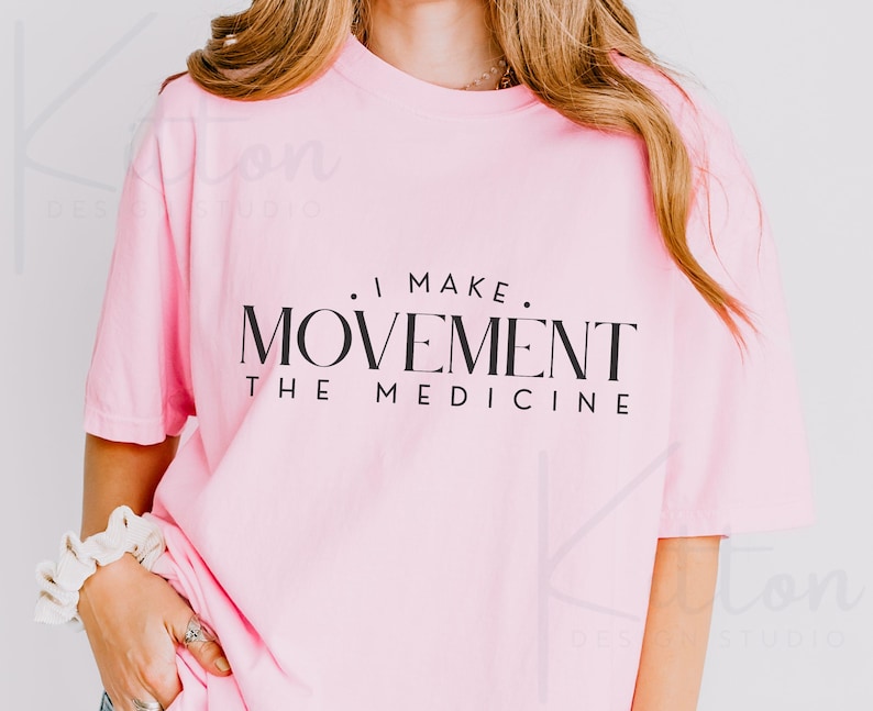 May include: A light pink t-shirt with the text "I MAKE MOVEMENT THE MEDICINE" in black. The shirt has short sleeves and a relaxed fit. The image is a close-up shot of the shirt.