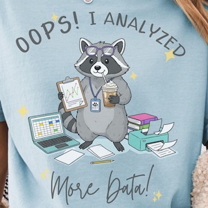 Puede incluir: Camiseta azul claro con un mapache de dibujos animados con gafas y sosteniendo una carpeta y un café. El texto "OOPS! I ANALYZED" está encima del mapache, y "More Data!" está debajo. Una computadora portátil, una impresora y papeles también están en la imagen.