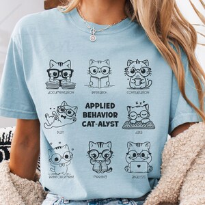 Puede incluir: Camiseta azul claro con un gráfico de gatos de dibujos animados con gafas, cada uno representando un aspecto diferente del Análisis de Conducta Aplicado. El texto de la camiseta dice "Applied Behavior Cat-alyst" con palabras como "Documentation" y "Research".