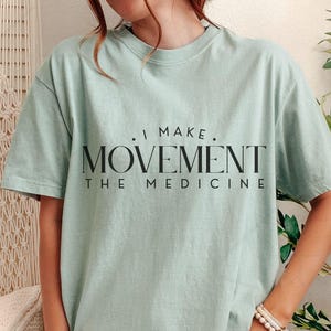 May include: A light green t-shirt with the text ". I MAKE. MOVEMENT THE MEDICINE" in black. The shirt has short sleeves and a crew neck. The shirt is made of a soft material.