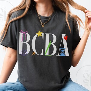 以下が含まれることがあります： 白い「BCBA」の文字が特徴の濃いグレーのTシャツ。各文字はカラフルなグラフィックで装飾されています。ピンクのリボン、黄色の星、緑のペン、赤いハート、青の砂時計がデザインに含まれています。シャツは柔らかい素材で作られています。