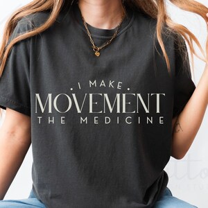 May include: Dark gray t-shirt with the text "I MAKE MOVEMENT THE MEDICINE" in a serif font. The shirt has short sleeves and a crew neck. A gold necklace is visible.
