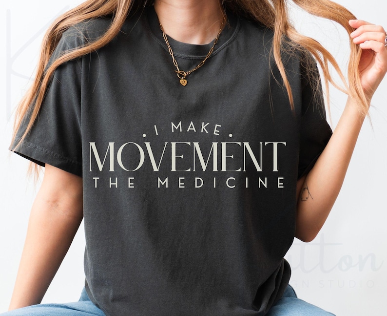 May include: Dark gray t-shirt with the text "I MAKE MOVEMENT THE MEDICINE" in a serif font. The shirt has short sleeves and a crew neck. A gold necklace is visible.