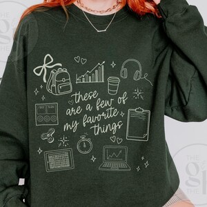 May include: Dark green sweatshirt with white line art illustrations of a backpack, coffee cup, headphones, laptop, and other objects. The text "these are a few of my favorite things" is also featured on the sweatshirt.