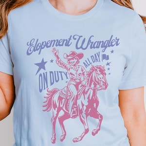 May include: Light blue t-shirt with a pink graphic of a cowgirl riding a horse, with the text "Elopement Wrangler ON DUTY ALL DAY". The design includes stars and a flag icon.