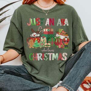 May include: An olive green t-shirt with the text "JUST AN ABA who loves CHRISTMAS" in a colorful, festive font. The shirt features Christmas-themed graphics, including a mug, gifts, a stocking, and a nutcracker.