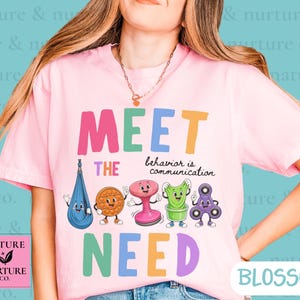 Op de afbeelding: Een lichtroze T-shirt met korte mouwen en de tekst MEET THE NEED en behavior is communication. De afbeelding bevat cartoonillustraties van een blauwe sensorische schommel, een oranje getextureerde schijf, een roze krukje, groene klei en een paarse fidget spinner.