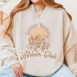 Op de afbeelding: Beige sweatshirt met een western thema. De afbeelding toont een cowgirl te paard met een lasso, met de woorden "Elopement Wrangler Athletic Club" in een vintage lettertype. De sweatshirt heeft een relaxte pasvorm.