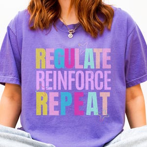 Peut inclure: T-shirt violet avec l'inscription "REGULATE REINFORCE REPEAT" en lettres multicolores. Le texte est en jaune, turquoise, rose et violet. Le t-shirt a des manches courtes et un col rond.
