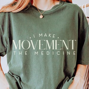 May include: Dark green t-shirt with the text "I MAKE MOVEMENT THE MEDICINE" in a cream-colored font. The shirt has a relaxed fit and short sleeves. The image shows the shirt on a person.