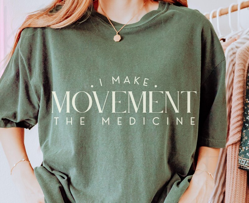 May include: Dark green t-shirt with the text "I MAKE MOVEMENT THE MEDICINE" in a cream-colored font. The shirt has a relaxed fit and short sleeves. The image shows the shirt on a person.