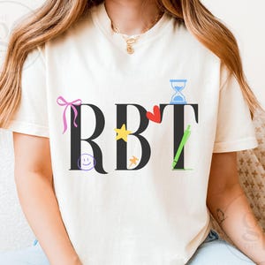 Puede incluir: Camiseta color crema con las letras negras "RBT". Las letras están decoradas con un lazo rosa, una carita sonriente, una estrella amarilla, un corazón rojo, un reloj de arena y un bolígrafo verde.