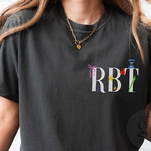 Puede incluir: Camiseta gris oscuro con las letras "RBT" en blanco, cada una adornada con gráficos coloridos. Un lazo rosa, una estrella amarilla, un corazón rojo y un tallo verde están entre los diseños. La camiseta tiene mangas cortas y cuello redondo.
