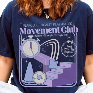May include: Navy blue t-shirt with a graphic design for a Movement Club. The design includes a stopwatch, a soccer ball, a cone, and the text "UNAPOLOGETICALLY PLAY-BASED Movement Club Building Strength Through Play Small Wins. Big Progress."