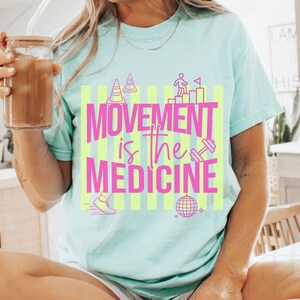 May include: A light blue t-shirt with the text "MOVEMENT is the MEDICINE" in pink and yellow. The graphic includes images of a person climbing stairs, a dumbbell, a disco ball, and a running shoe.