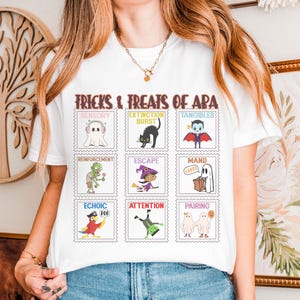 Puede incluir: Camiseta blanca con un diseño gráfico colorido. El texto dice "FRICKS & TREATS OF APA" y presenta ilustraciones temáticas de Halloween, incluyendo un fantasma, un gato negro, un vampiro, un zombi, una bruja y otros personajes espeluznantes.