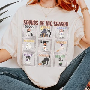 Può includere: T-shirt color crema con un design a tema Halloween. La maglietta mostra una serie di immagini simili a francobolli con fantasmi, una casa infestata e un gatto nero, insieme al testo "SOUNDS OF THE SEASON".