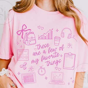 May include: Pink t-shirt with a graphic design in a lighter shade of pink. The design includes a backpack, hearts, a graph, headphones, a coffee cup, a clipboard, a timer, a laptop, and the text "these are a few of my favorite things."