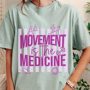 May include: A light green t-shirt with the text "MOVEMENT is the MEDICINE" in bright pink. The design includes illustrations of traffic cones, a person climbing stairs, a dumbbell, a running shoe, and a disco ball.
