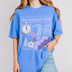May include: A light blue t-shirt with a graphic design for the "Movement Club." The design includes a stopwatch, a soccer ball, a traffic cone, and the text "Unapologetically Play-Based." The shirt also has the text "Building Strength Through Play."