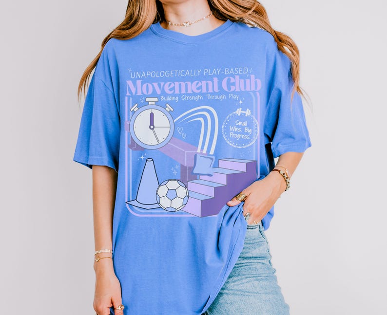 May include: A light blue t-shirt with a graphic design for the "Movement Club." The design includes a stopwatch, a soccer ball, a traffic cone, and the text "Unapologetically Play-Based." The shirt also has the text "Building Strength Through Play."