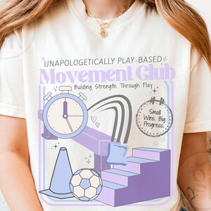 May include: Cream-colored t-shirt featuring a graphic design with the text "UNAPOLOGETICALLY PLAY-BASED Movement Club." The design includes a stopwatch, a soccer ball, a cone, and a set of stairs in shades of purple and blue. The phrase "Small Wins. Big Progress." is also included.