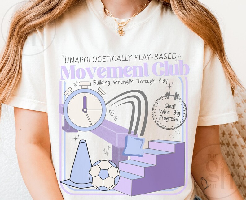 May include: Cream-colored t-shirt featuring a graphic design with the text "UNAPOLOGETICALLY PLAY-BASED Movement Club." The design includes a stopwatch, a soccer ball, a cone, and a set of stairs in shades of purple and blue. The phrase "Small Wins. Big Progress." is also included.