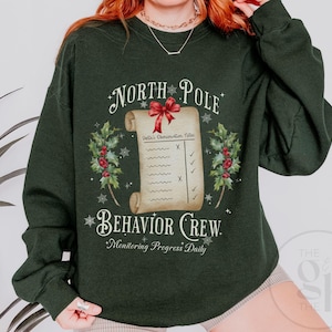 Op de afbeelding: Donkergroene sweatshirt met een feestelijk ontwerp. De afbeelding toont de tekst "North Pole Behavior Crew" rond een rol met een rode strik en hulstaccenten. De rol leest "Santa's Observation Notes".